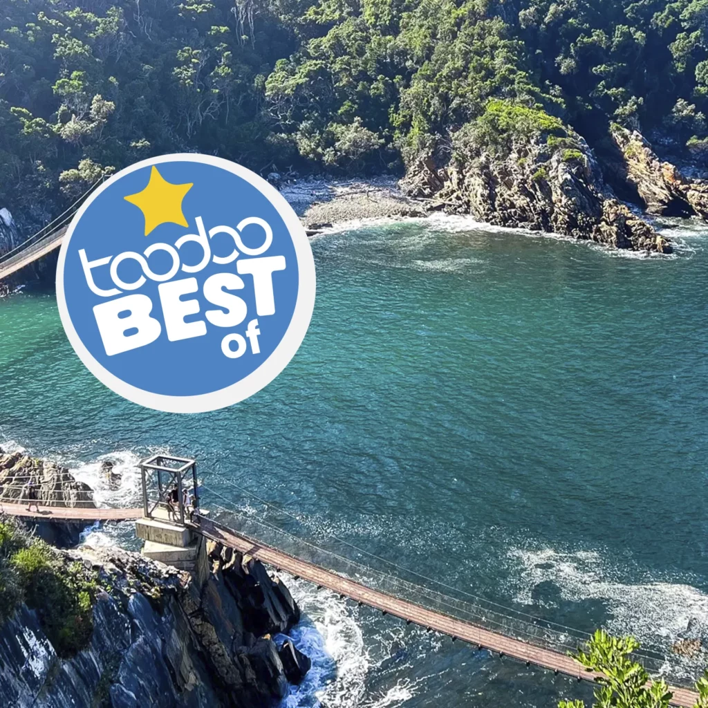 Suspension bridge over rocky coastline and blue waters in the Garden Route, South Africa, with a toodoo "Best Of" badge.